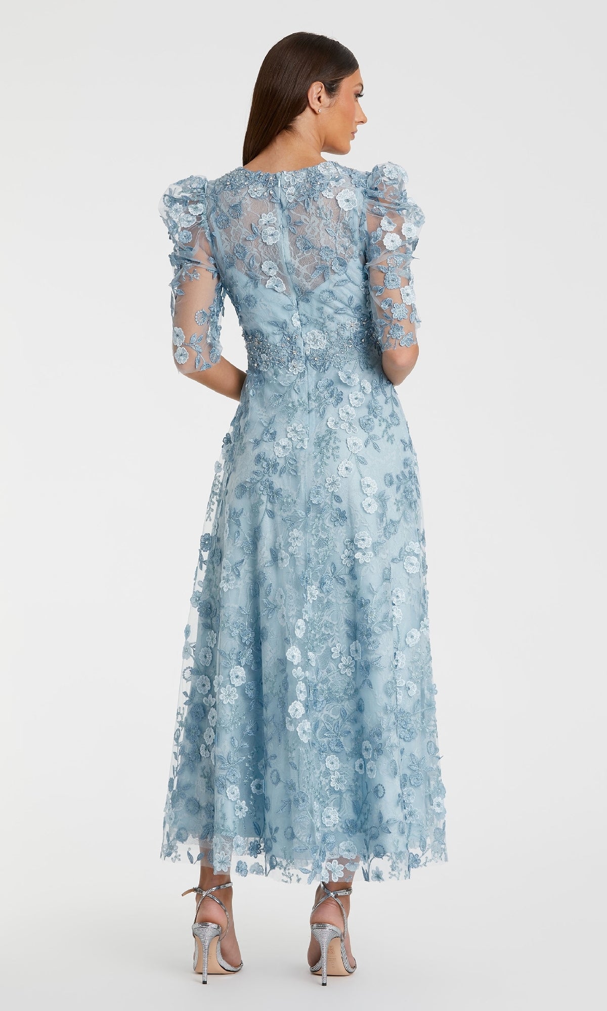 A woman stands facing away, wearing the Mac Duggal 68193 tea-length lace formal dress with floral patterns, puffed short sleeves, and silver high-heeled sandals.